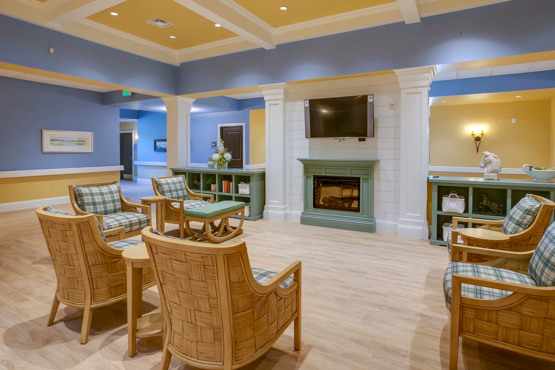 A lounge area with wicker armchairs and plaid cushions arranged around a low table facing a wall-mounted TV above a fireplace.