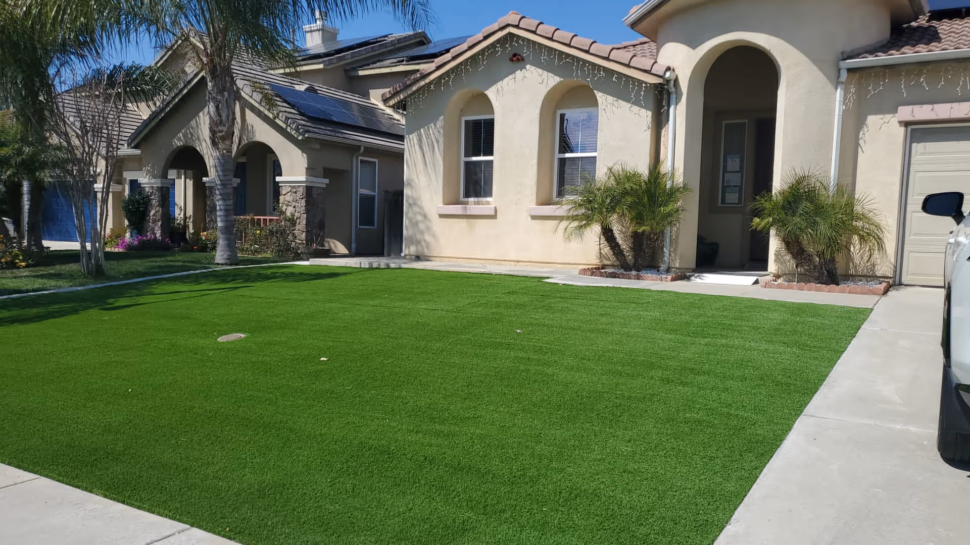 Front exterior view of a single-story beige house with a well-maintained green lawn, palm trees, and a driveway with a white vehicle partially visible on the right side.