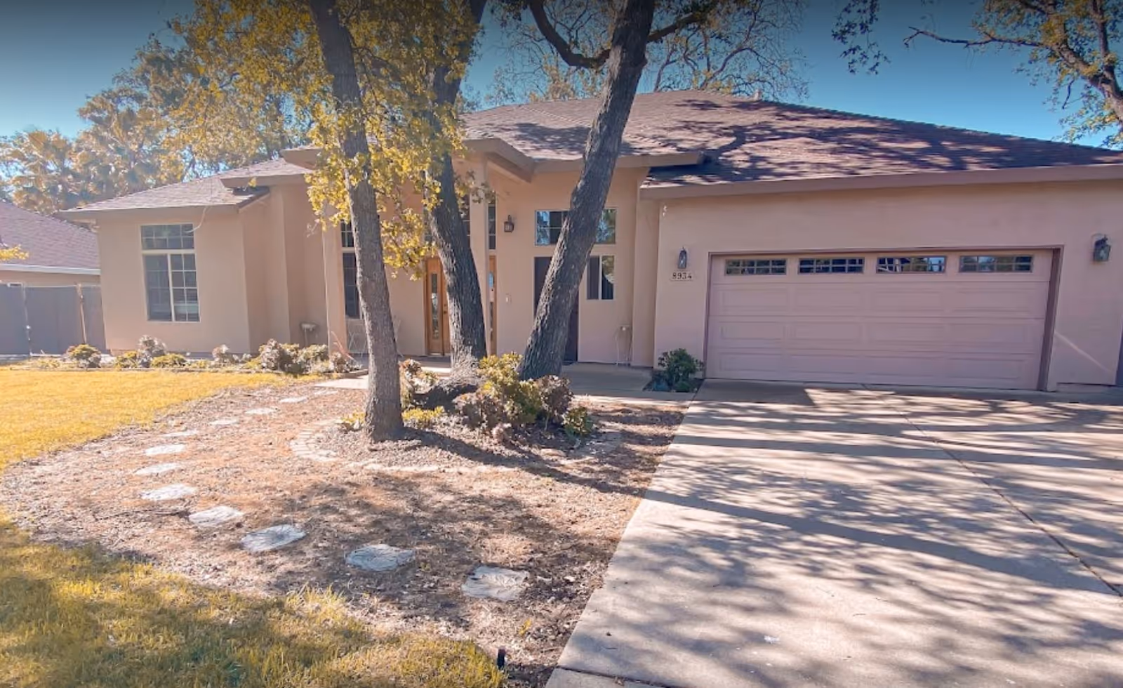 Front exterior view of a single-story residential building with a two-car garage, a driveway, and a small garden area with trees and stepping stones leading to the entrance.