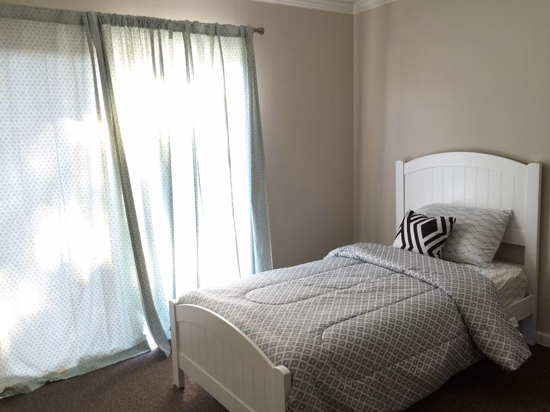 A small bedroom with a single white wooden bed against a beige wall. The bed is made with a gray and white patterned comforter, a pillow with a similar pattern, and a black and white geometric accent pillow. To the left, there is a large window covered by light blue patterned curtains with sunlight filtering through.
