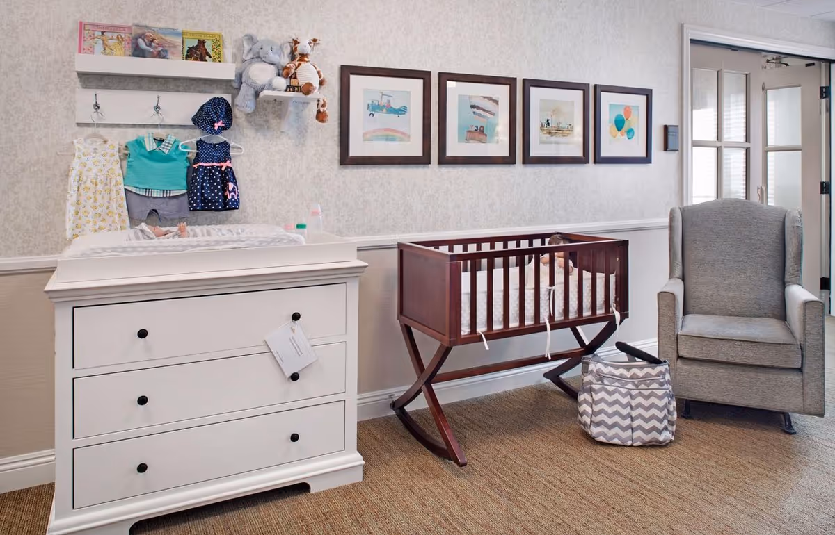 A nursery-style interior with a white changing dresser, a wooden crib, gray armchair, and framed artwork on the wall.