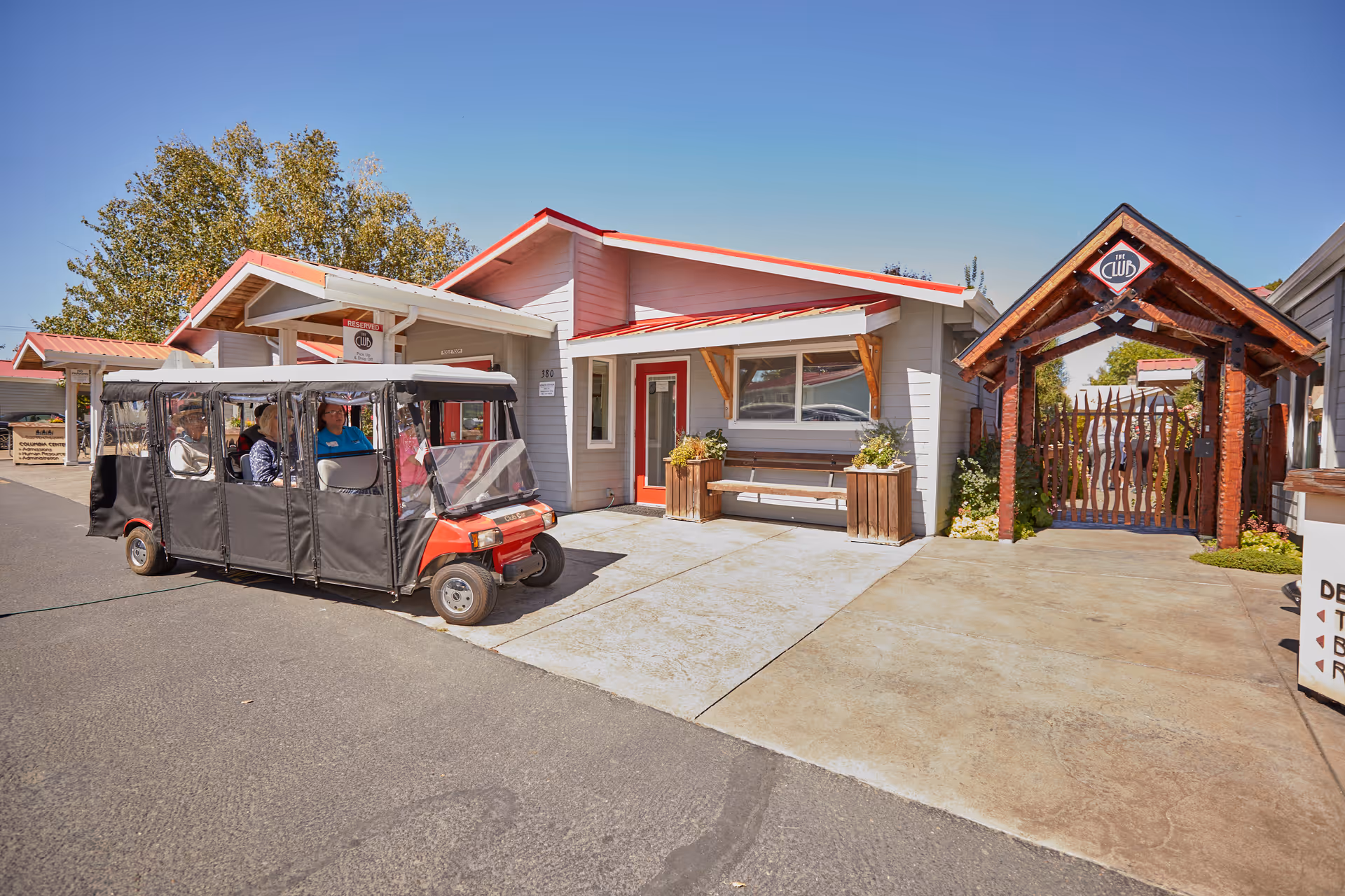 Exterior view of a senior living facility building with a red roof and gray walls. A covered golf cart with passengers is parked on the driveway. To the right, there is a wooden gate entrance with a sign that reads 'The Club'. Trees and clear blue sky are visible in the background.