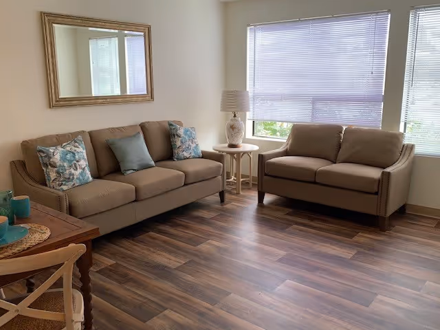 A living room with two beige sofas adorned with decorative pillows, a wooden side table with a lamp, a large window with blinds, a wooden floor, and a framed mirror on the wall.