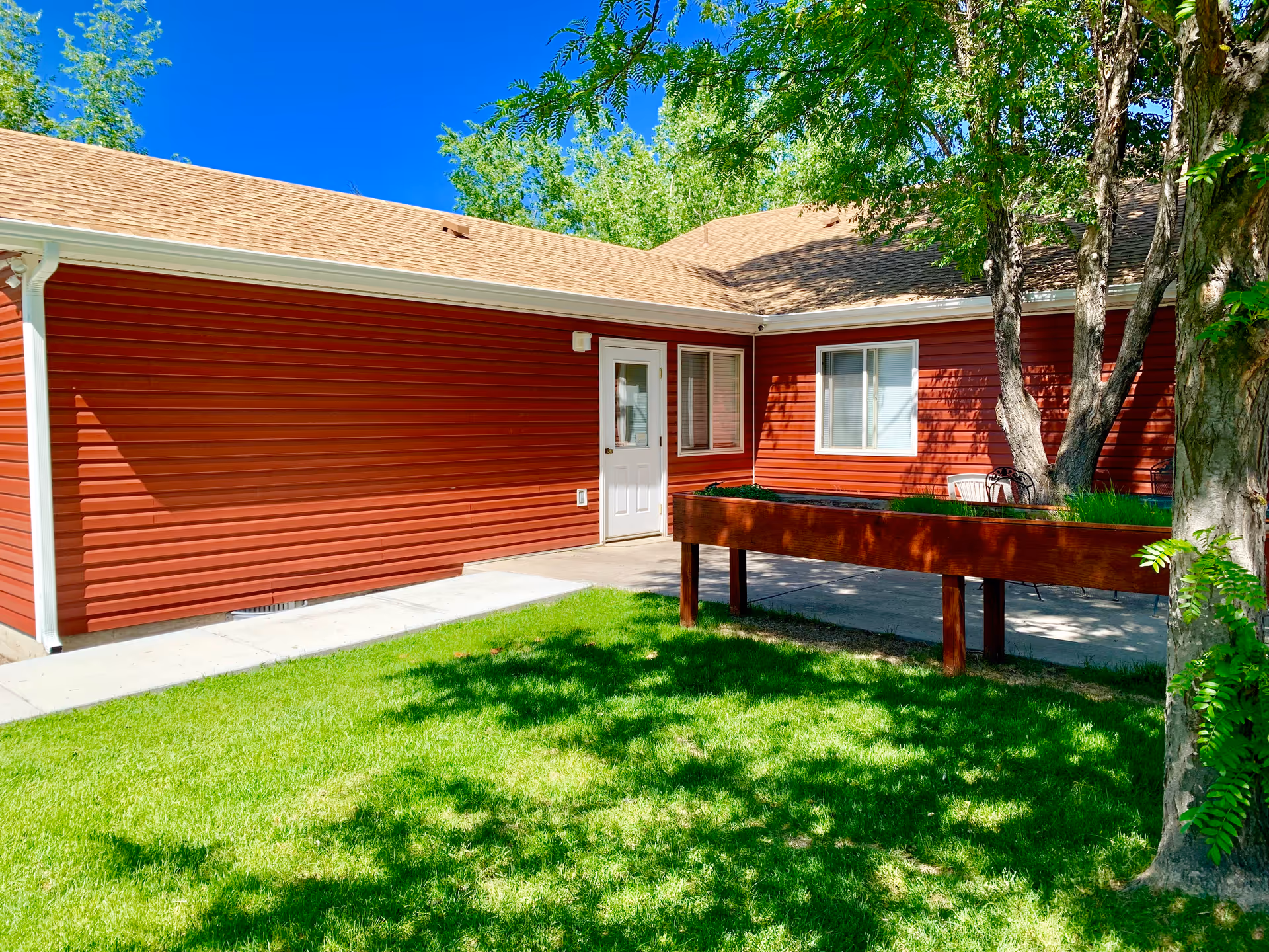 Outdoor view of a red building with a beige roof, a white door, and windows. There is a green lawn in the foreground with shadows from nearby trees, and a raised wooden planter box near the building. Trees and clear blue sky are visible in the background.