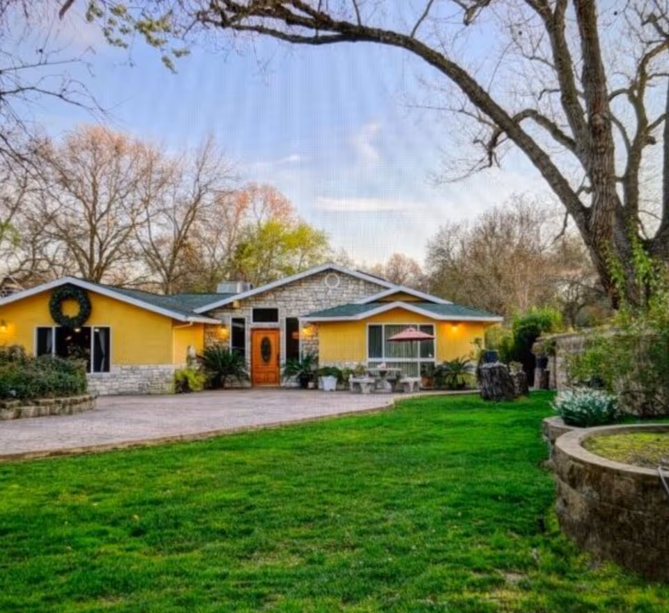 Front exterior view of a single-story yellow building with stone accents, a wooden door, and a wreath on one window. The building is surrounded by a green lawn, trees with bare branches, and outdoor seating with an umbrella.