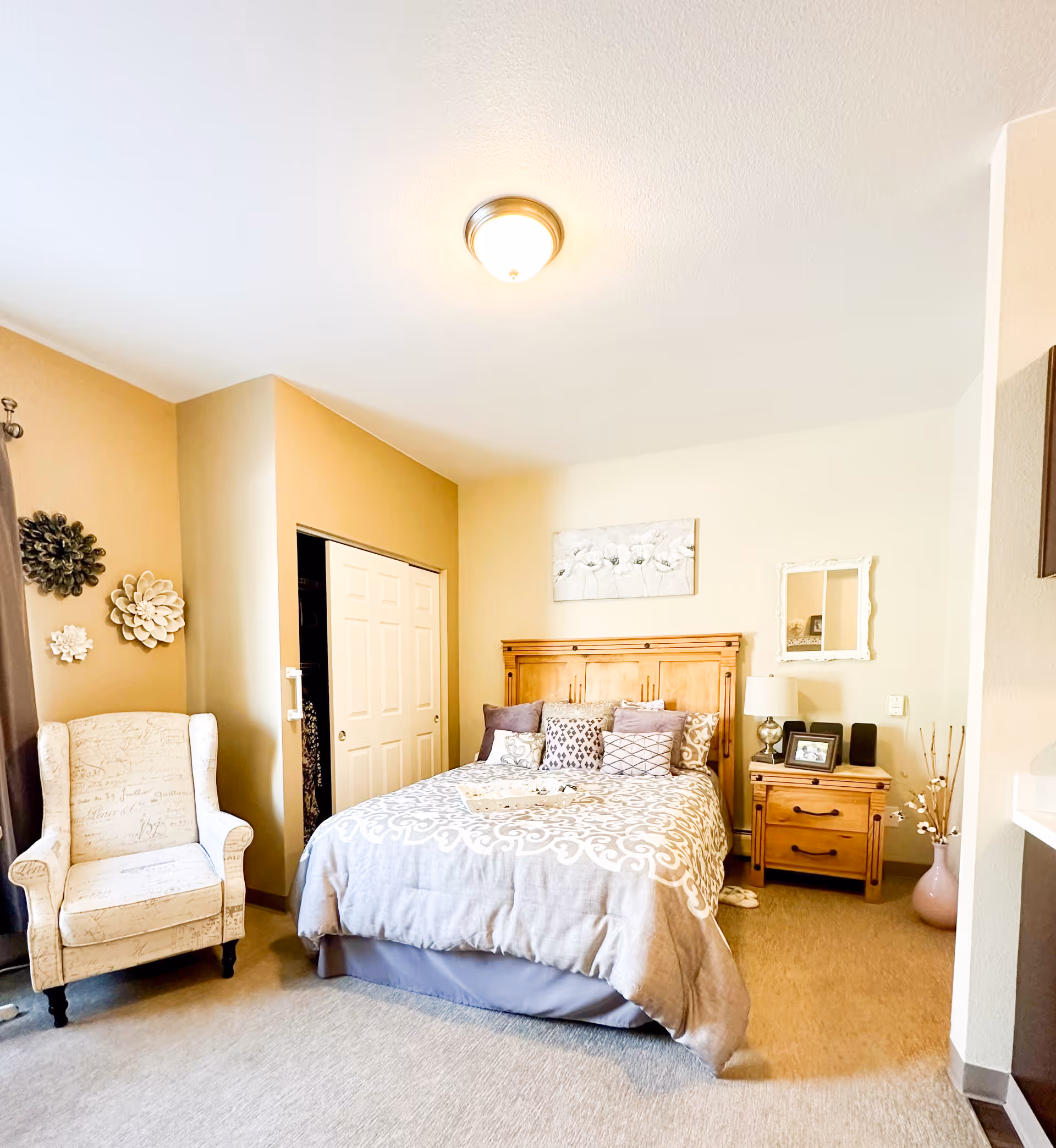 Sunny, warmly decorated bedroom with a wooden headboard, patterned bedding, a nightstand, and an upholstered armchair.