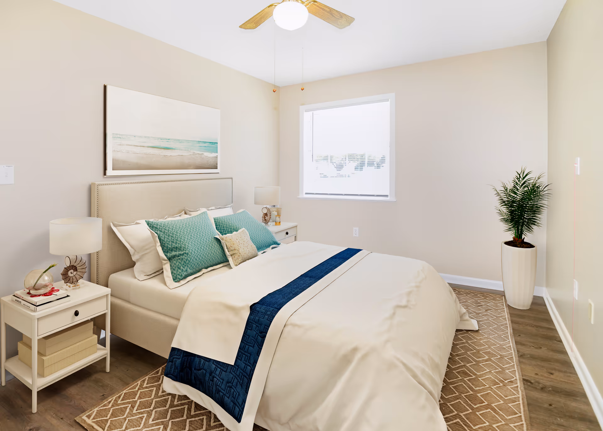 A neatly made bed with white and teal bedding in a bright bedroom. The room features a beige upholstered headboard, two white nightstands with lamps and decorative items, a large window with blinds, a ceiling fan with wooden blades, a patterned area rug, and a tall potted plant in the corner.