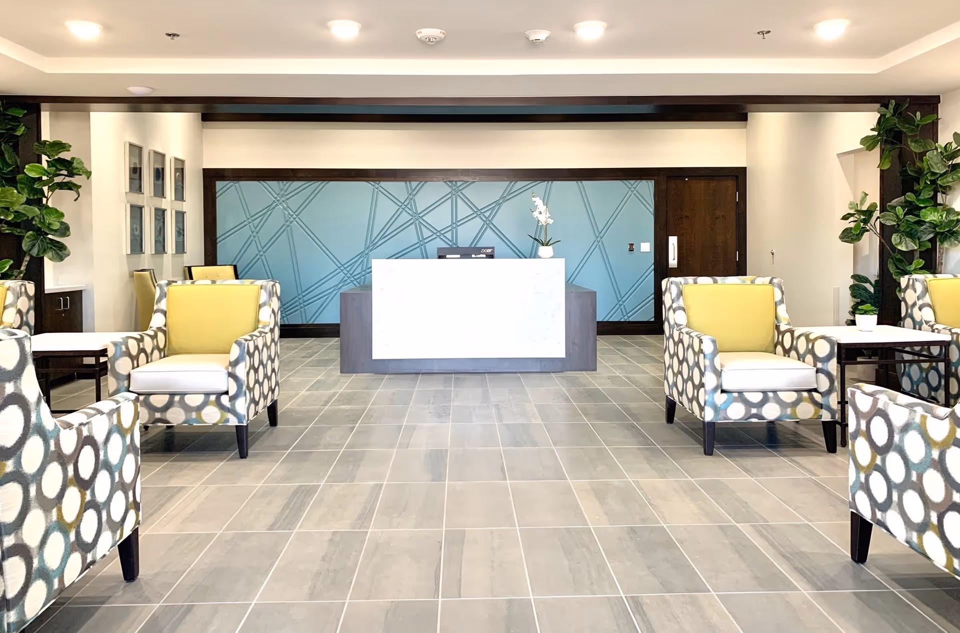 A modern reception area with a white front desk against a blue wall featuring a geometric pattern. The room has tiled flooring and is furnished with patterned armchairs with yellow cushions, small tables, and green plants on either side.