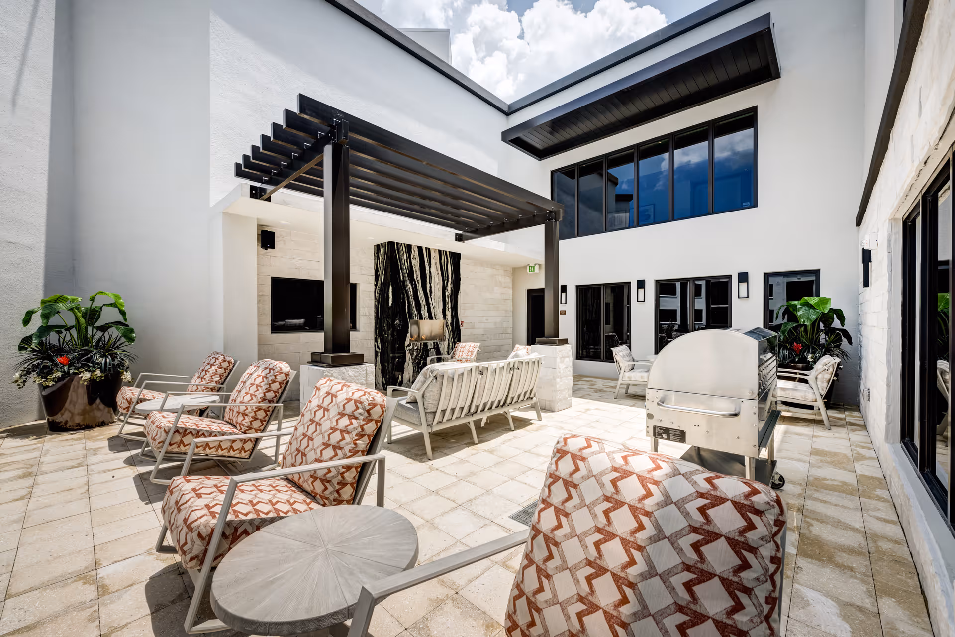 Outdoor courtyard patio with cushioned lounge chairs, a pergola-covered seating area, a grill, and potted plants.