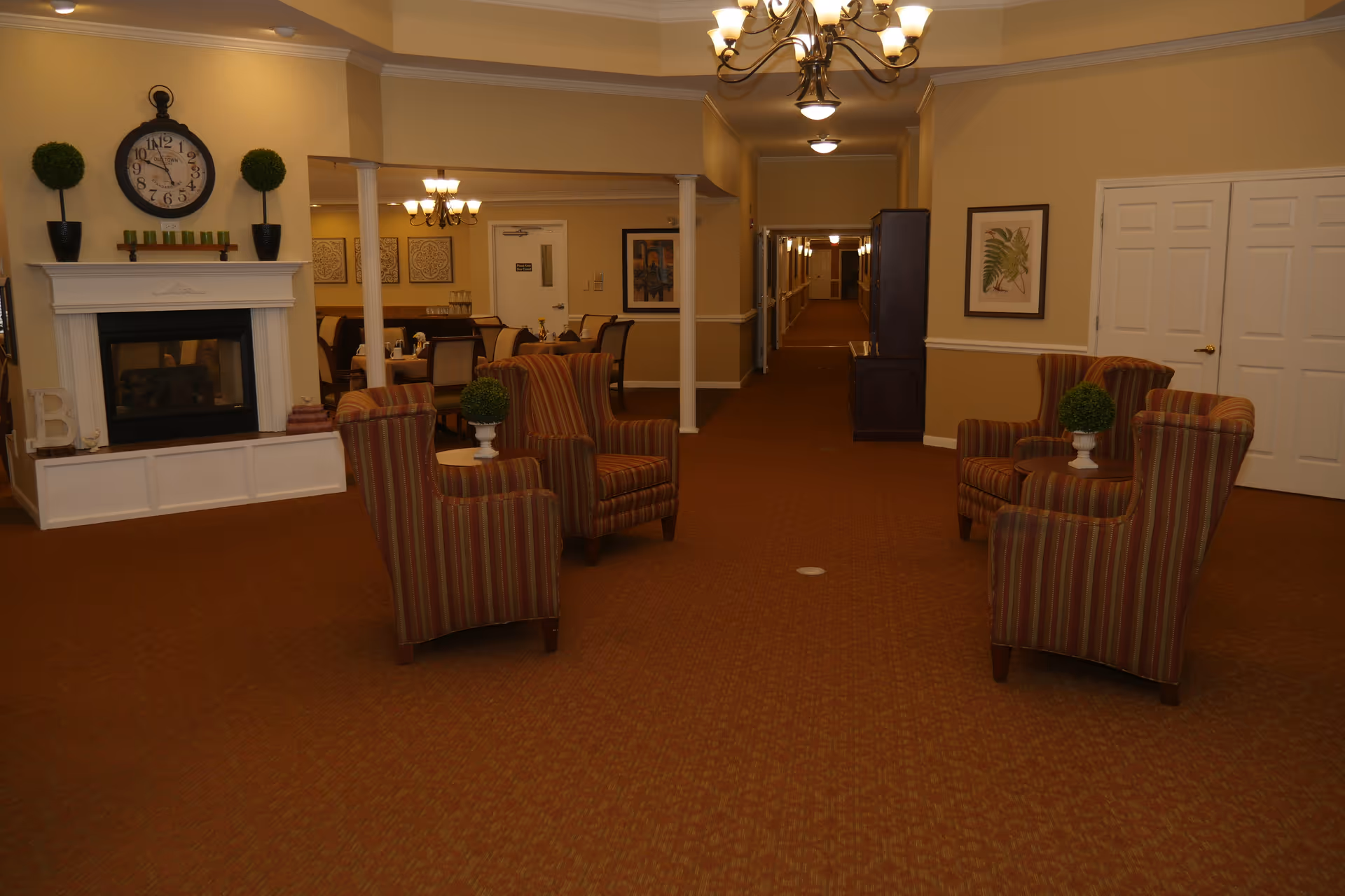 A warm interior lounge with striped armchairs arranged around small tables, a fireplace, chandeliers, and a long hallway leading to dining areas.