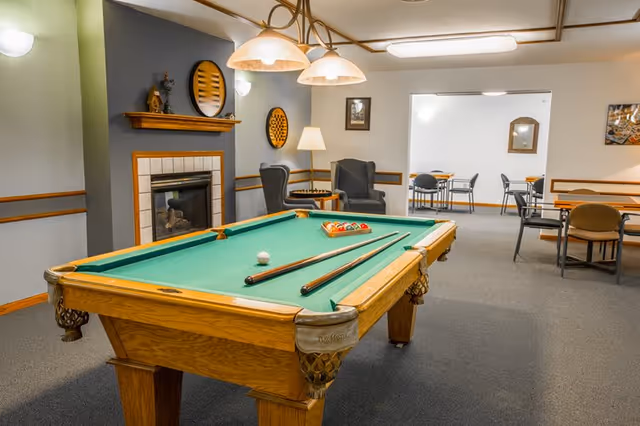 A recreational room with a pool table in the center, two pool cues and a rack of billiard balls on the table. The room features a fireplace with decorative items on the mantel, two armchairs with a floor lamp between them, and several tables and chairs in an adjoining room visible through an open doorway. The walls are painted in neutral tones with wood trim, and overhead lighting fixtures hang above the pool table.