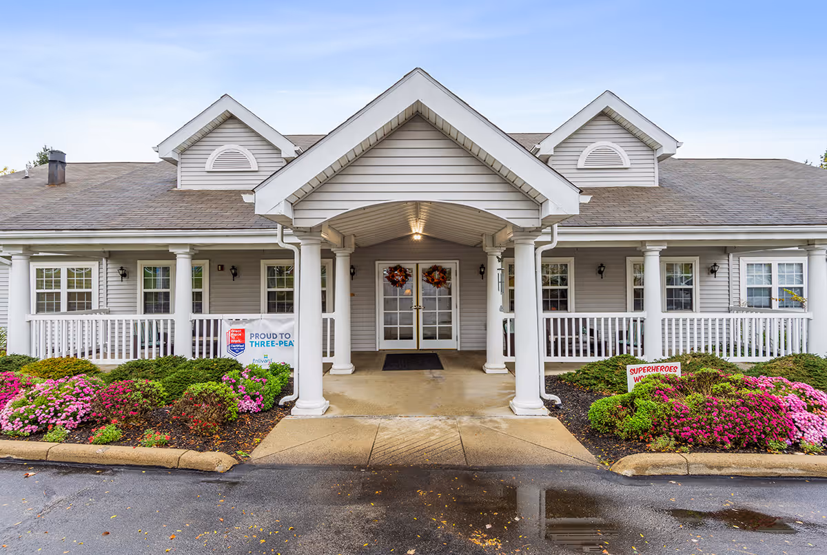 Front exterior view of a single-story building with a covered entrance supported by white columns. The building has gray siding, multiple windows, and a porch with white railings. There are colorful flower beds with pink and red flowers on either side of the entrance. Two wreaths hang on the glass double doors at the entrance.