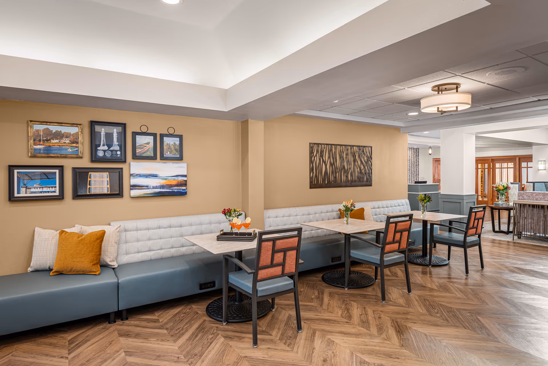 A cozy dining area in Harbour Village featuring a long cushioned bench along a beige wall adorned with framed artwork. There are three small tables with chairs, each table decorated with a small vase of flowers. The floor has a wood-patterned design, and the ceiling has modern light fixtures.