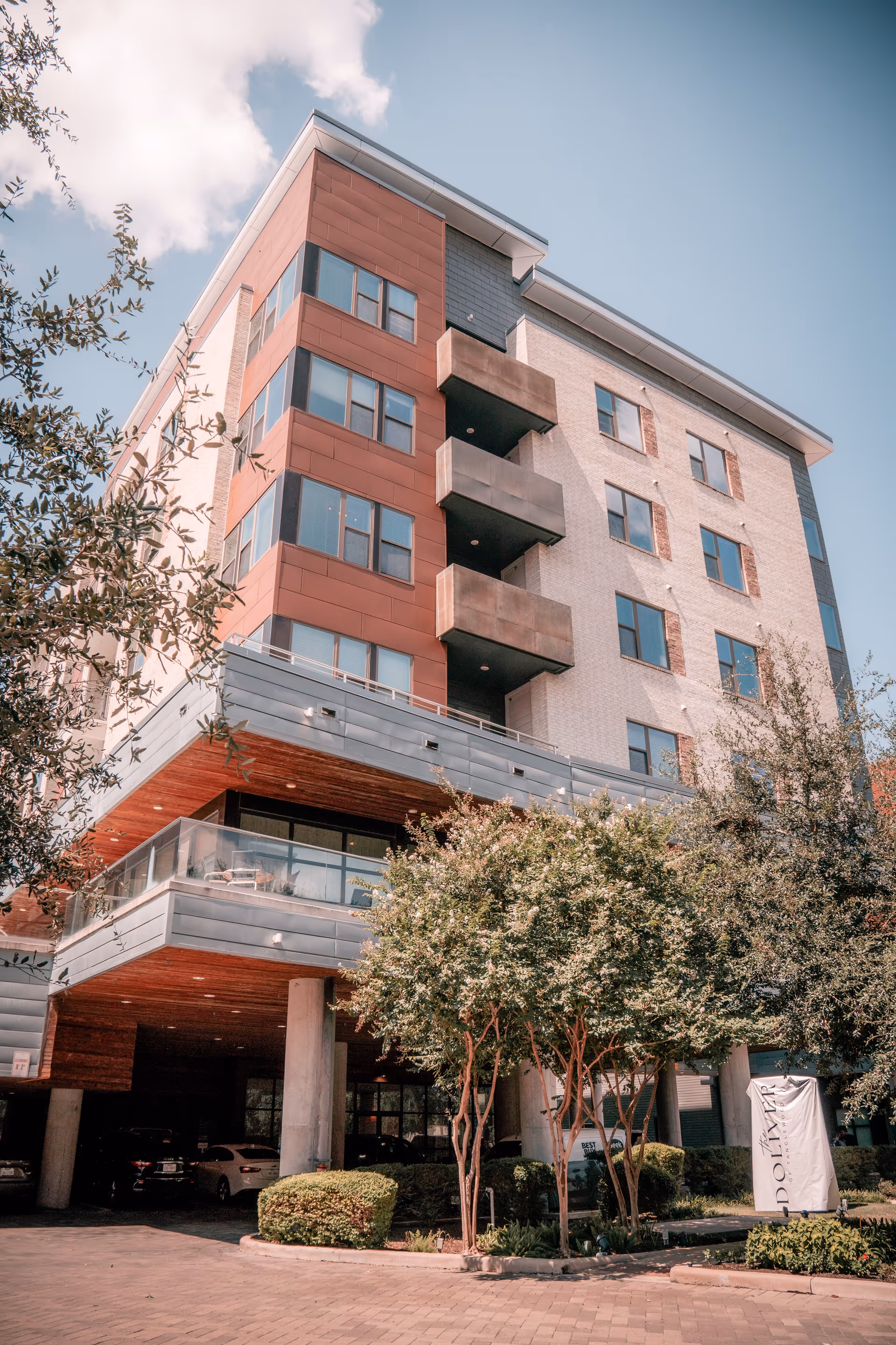 Exterior view of a modern multi-story senior living facility building with a combination of brick and panel siding, balconies, and large windows. The building is surrounded by trees and landscaping, with a covered parking area underneath. A sign near the entrance reads 'Doliver'.