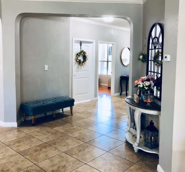 A bright and clean interior hallway with beige tiled floors and light gray walls. There is a teal cushioned bench on the left side and a white console table with flowers, a clock, and decorative items on the right. A large decorative mirror hangs above the console table. Two white doors are visible at the end of the hallway, one adorned with a wreath. Natural light streams in from a window in an adjoining room.