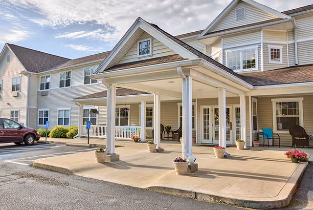 Exterior view of a senior living facility building with a covered entrance supported by white columns. There are several potted plants with flowers placed around the entrance area. The building has beige siding, multiple windows, and a brown shingled roof. A maroon car is parked in a parking space nearby, and the sky is partly cloudy.