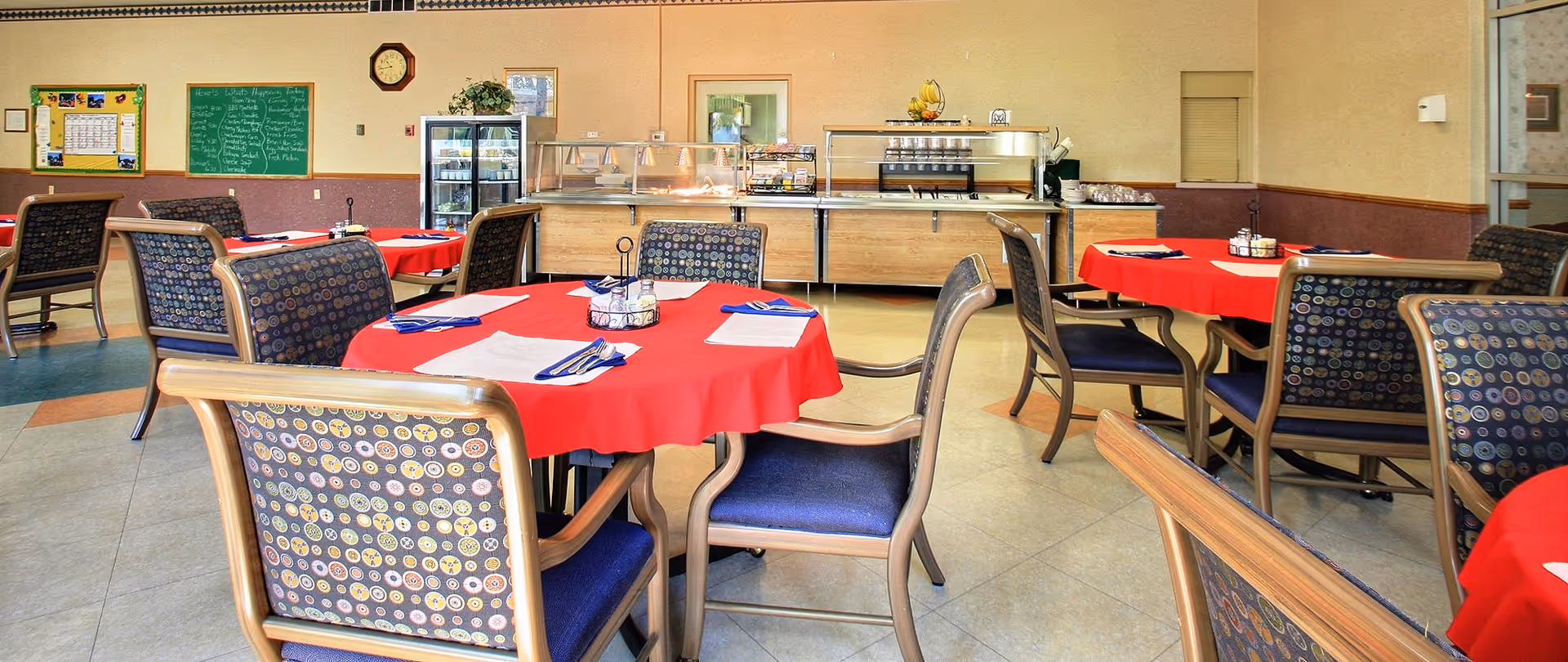 A dining room in a senior living facility with round tables covered in red tablecloths. Each table is set with napkins and utensils, and surrounded by wooden chairs with patterned cushions. In the background, there is a food serving area with a glass sneeze guard, a refrigerator, a chalkboard menu, and a clock on the wall.