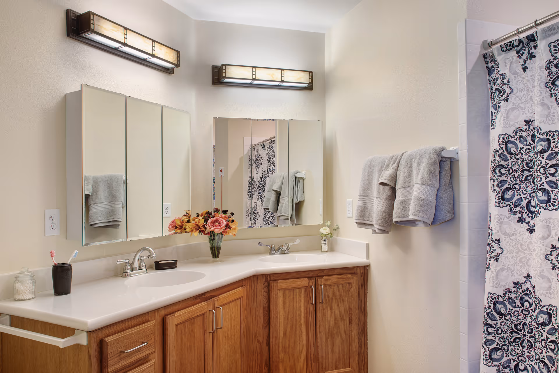 Bright bathroom with a double-sink white countertop over wooden cabinets, wall-mounted mirrors and lights, folded towels, a vase of flowers, and a patterned shower curtain.