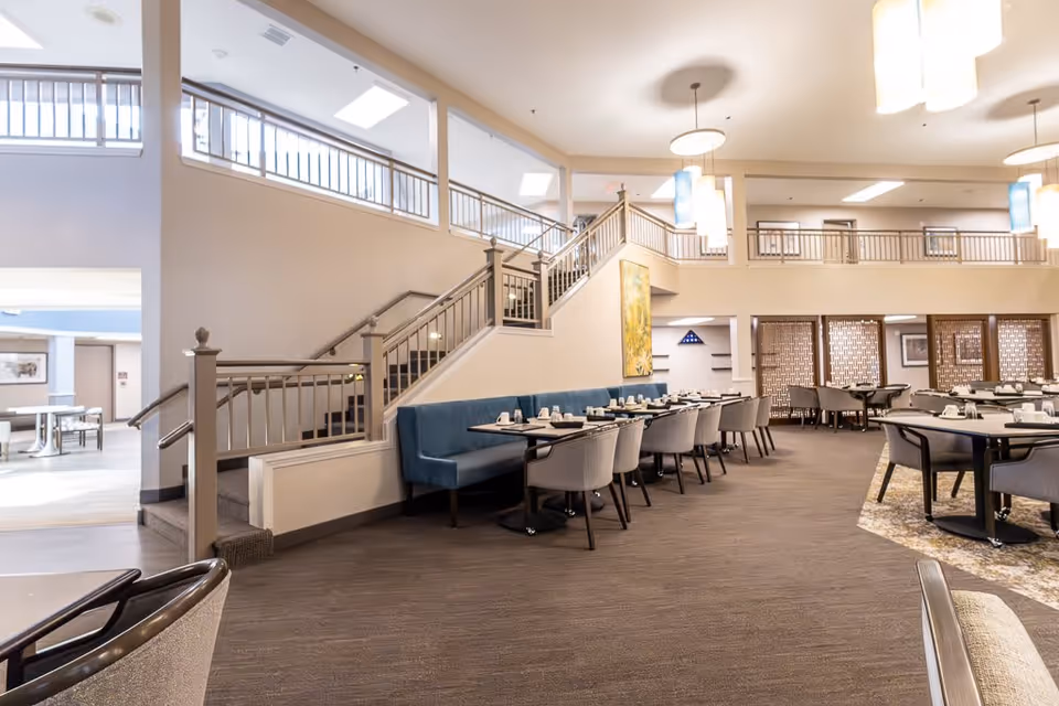 Spacious dining area with multiple tables and chairs arranged neatly. There is a blue cushioned bench along the wall under a staircase, and the room features modern hanging light fixtures and neutral-toned decor.