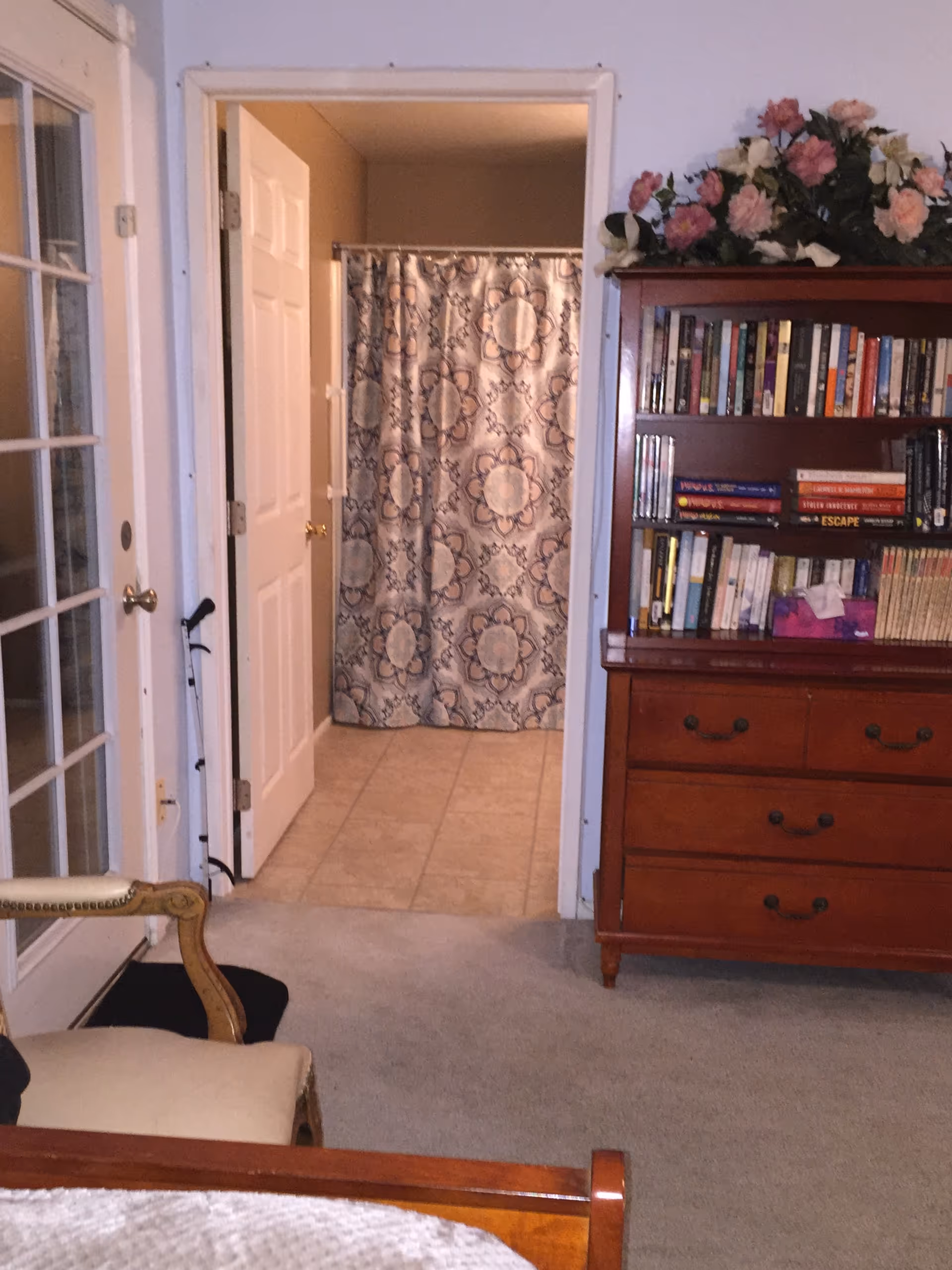 Interior view of a bedroom area with a wooden dresser and bookshelf, a chair, and a doorway leading to a tiled bathroom with a patterned shower curtain.