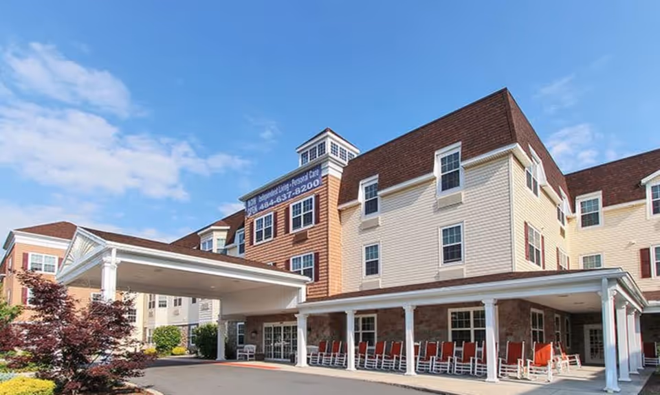 Exterior view of Keystone Villa at Fleetwood, a multi-story senior living facility with beige and brown siding, white trim, and a covered entrance. Red rocking chairs are lined up under the covered porch area. The sky is blue with some clouds.