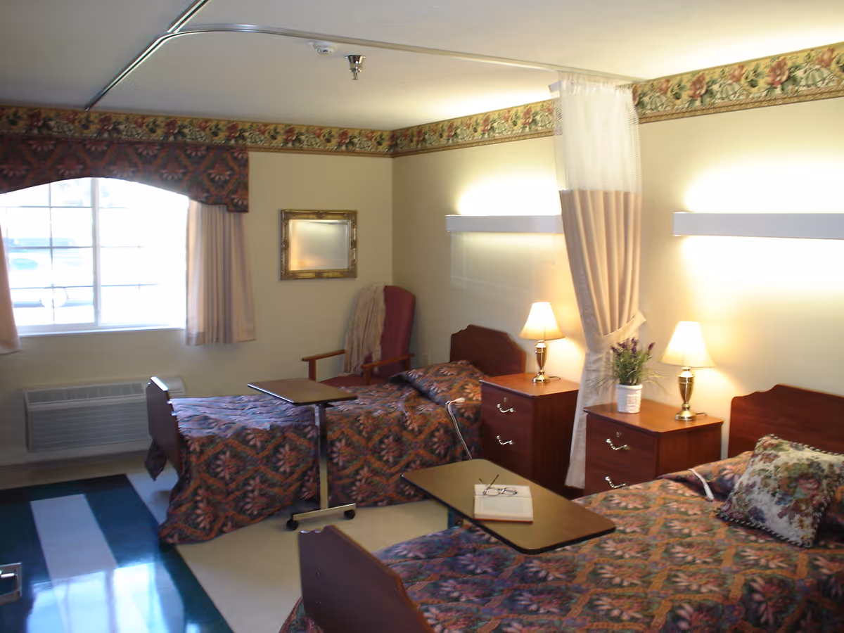 A shared bedroom in a senior living facility with two single beds covered in patterned bedspreads. Each bed has a bedside table with a lamp, and there is a small table on wheels positioned over each bed. A window with curtains and a valance is on the left wall, and a comfortable armchair with a throw blanket is placed in the corner. The room has a floral border near the ceiling and a curtain divider between the beds.