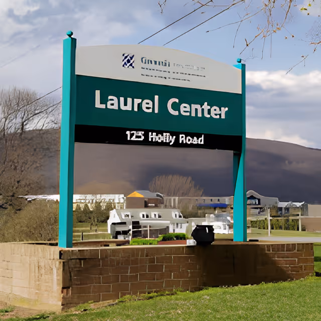 Green roadside sign reading 'Laurel Center' on teal posts with a grassy foreground and hills and buildings in the background.