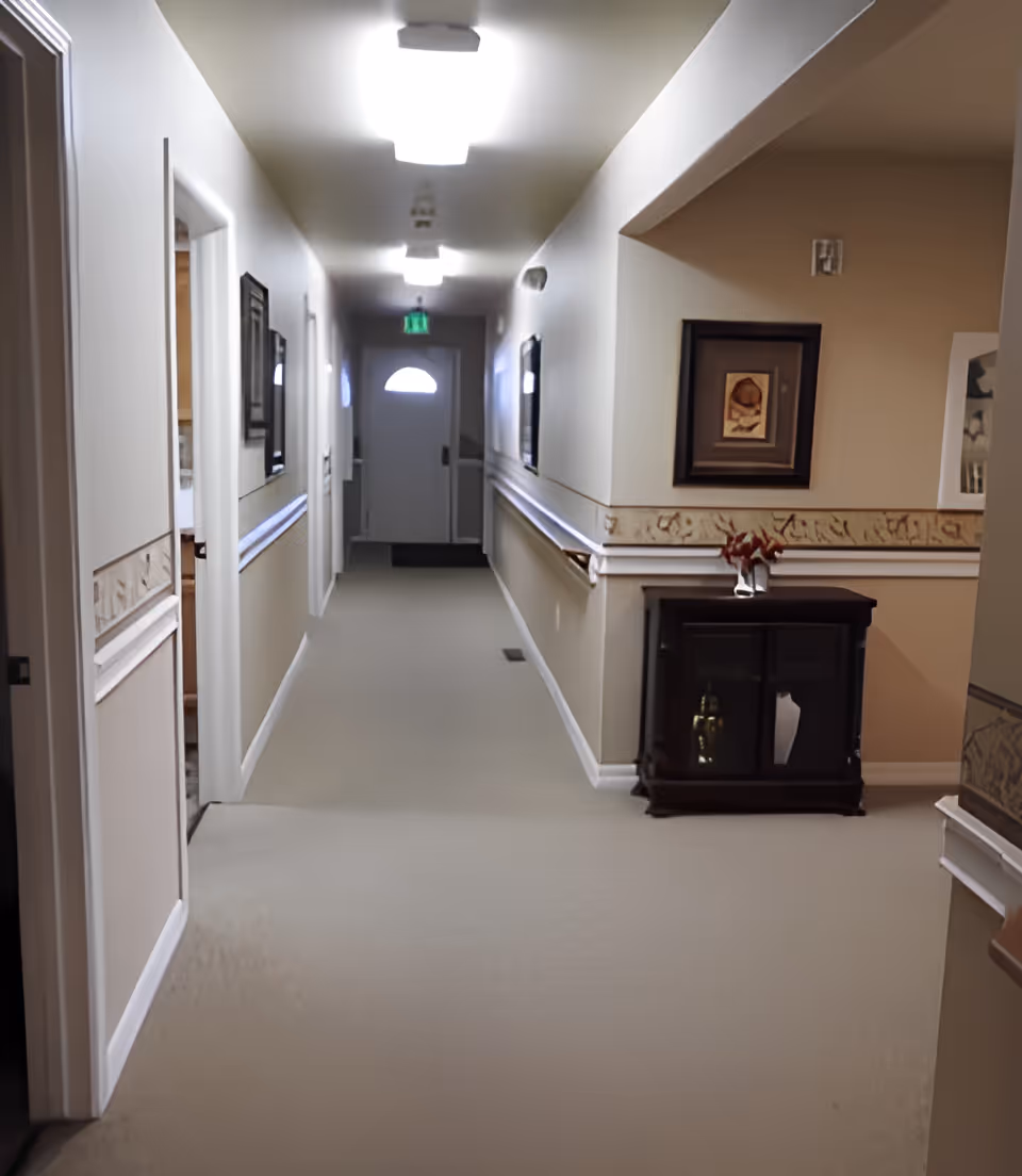 A well-lit interior hallway in an assisted living facility with carpeted floor, handrails, framed artwork, and a small cabinet.