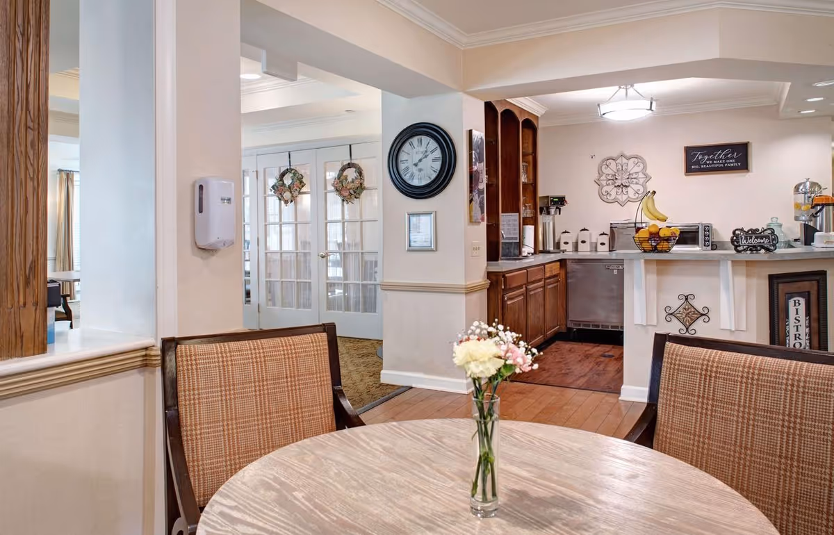 Interior view of a senior living facility dining area with a round wooden table and two cushioned chairs in the foreground. A small vase with flowers is on the table. In the background, there is a kitchen area with wooden cabinets, a countertop with a fruit basket, coffee machine, and other kitchen items. A wall clock and decorative signs are visible on the walls, along with glass double doors adorned with wreaths.