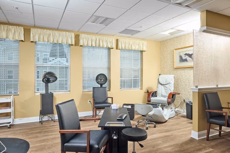 Interior view of a salon area in a senior living facility with multiple salon chairs, hair dryers, a pedicure spa chair, and a manicure table. The room has beige walls, three windows with blinds and yellow valances, and wood flooring.