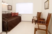A small, simple room with a brown daybed against the left wall, adorned with a red patterned pillow. Two wooden chairs with beige cushions are placed against the right wall. A large window with a white blind is centered on the back wall. The walls are light-colored, and there are two framed pictures hanging on the walls. The floor is carpeted in a light color.