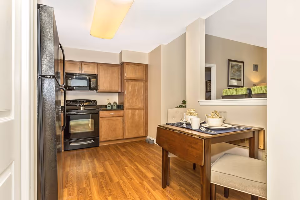 Small open kitchen with wooden cabinets, black appliances and a small dining table set for two.
