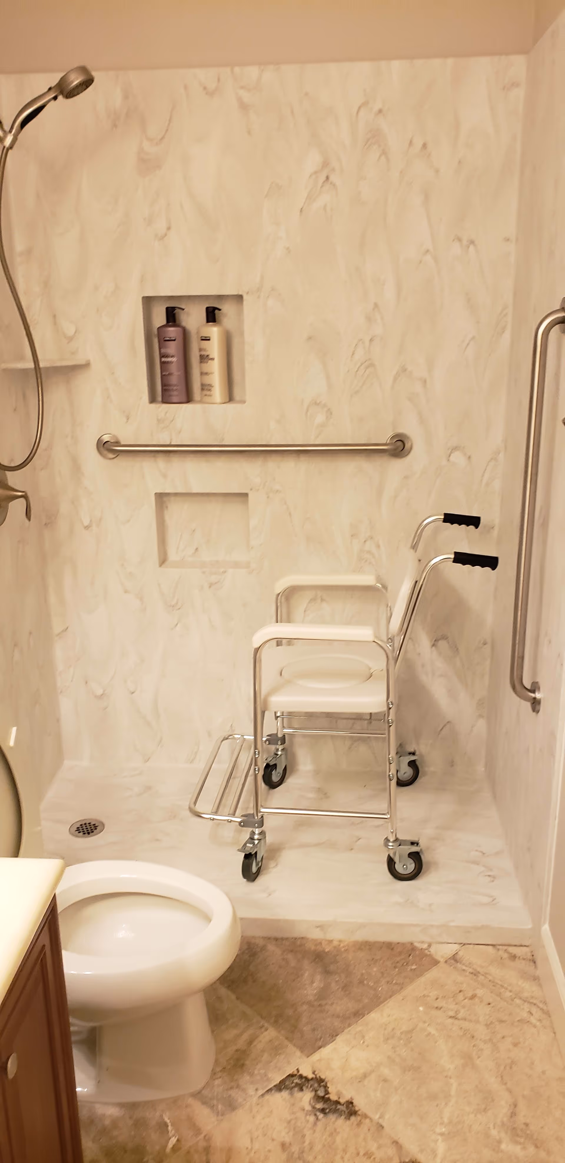 A bathroom with a toilet, a walk-in shower equipped with a handheld showerhead, grab bars, and a shower chair with wheels. There are two bottles of shampoo or body wash placed in a recessed shelf in the shower wall.