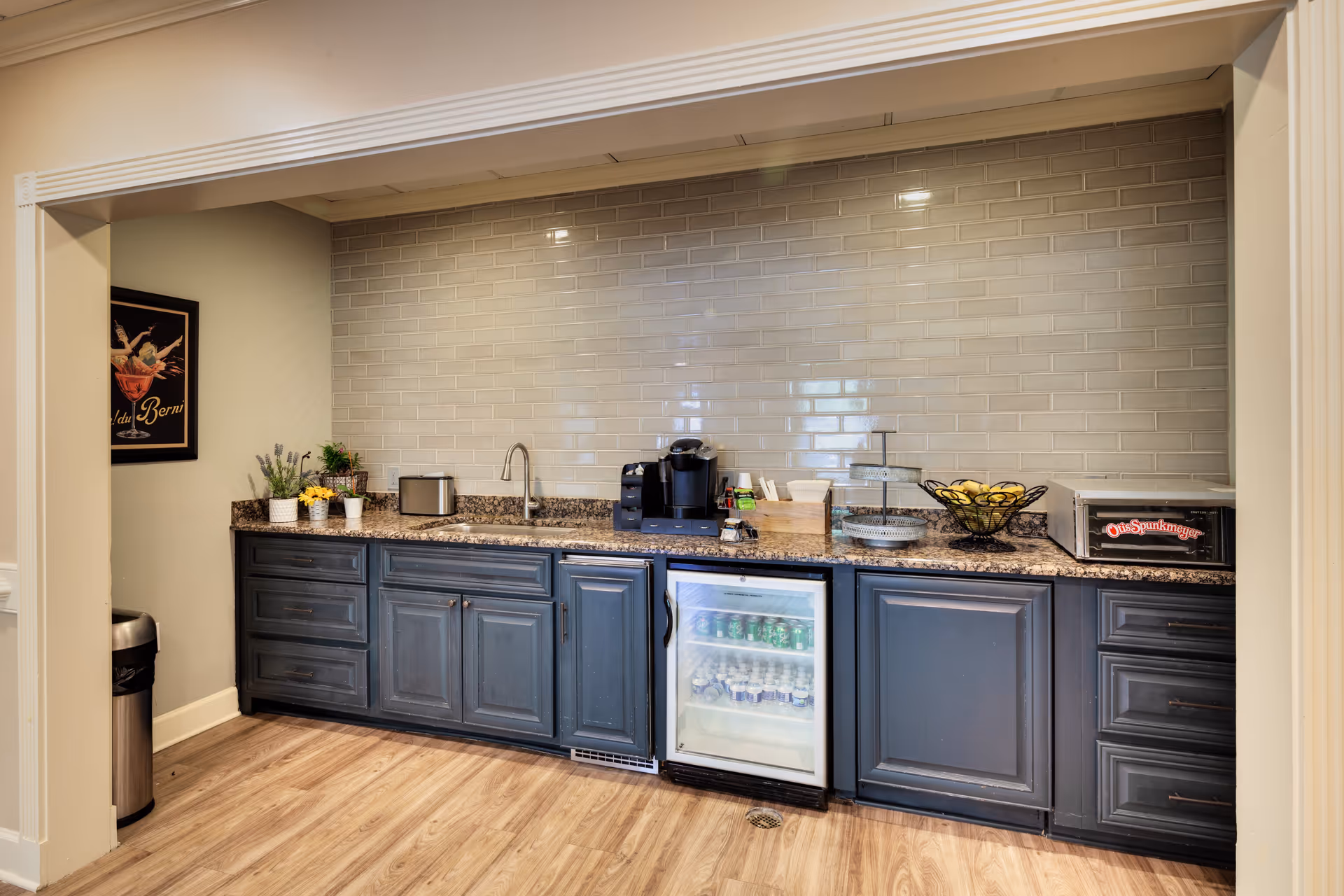 A kitchenette area with dark blue cabinets, a granite countertop, a small sink, a coffee maker, a mini fridge stocked with beverages, a toaster oven, and a bowl of lemons. The backsplash is made of glossy beige subway tiles, and the floor is light wood. There is a framed picture on the left wall and a stainless steel trash can below it.