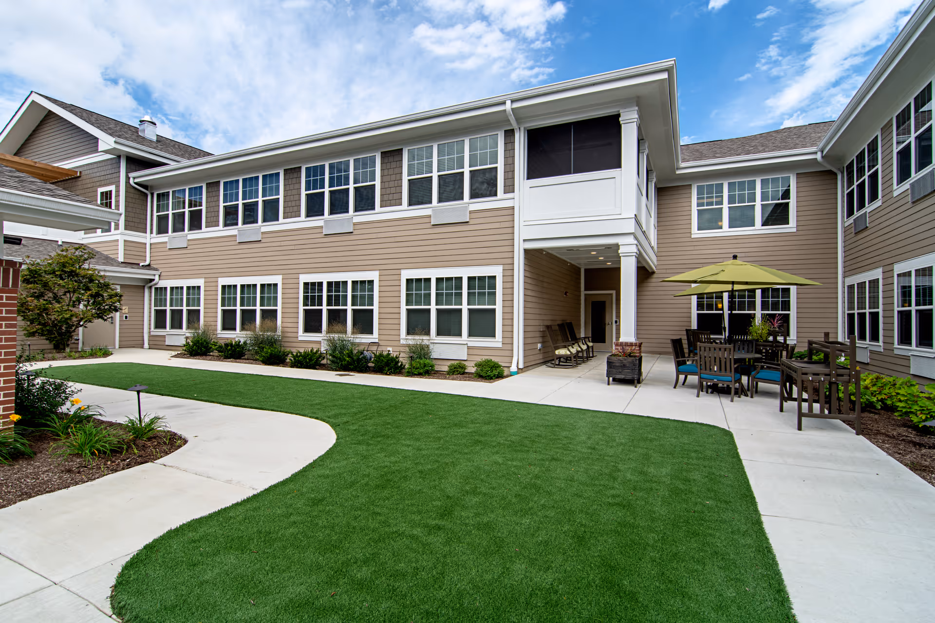 Courtyard of a two-story beige senior living building with a manicured lawn, concrete walkways and patio seating with an umbrella.