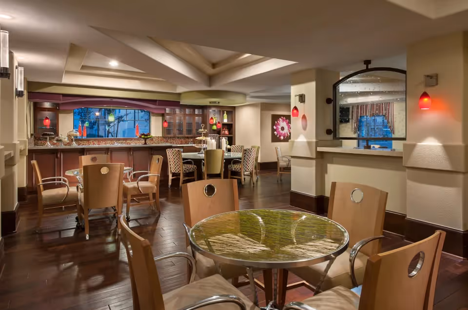 Interior view of a dining area in Clearwater Agritopia featuring round tables with wooden chairs, a counter with bar stools, pendant lights with red shades, and a large window in the background.