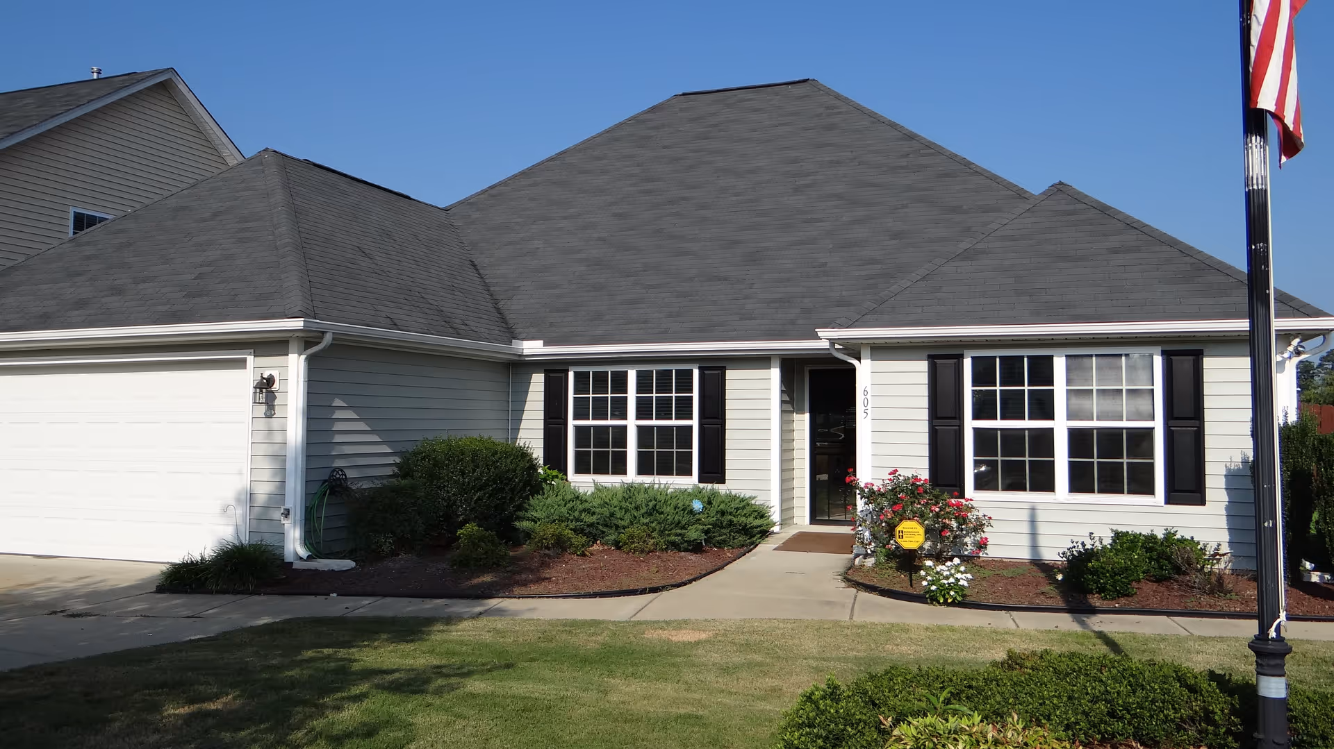 Single-story light-gray house front with an attached garage, black-shuttered windows, walkway and landscaped beds with an American flag on a pole.