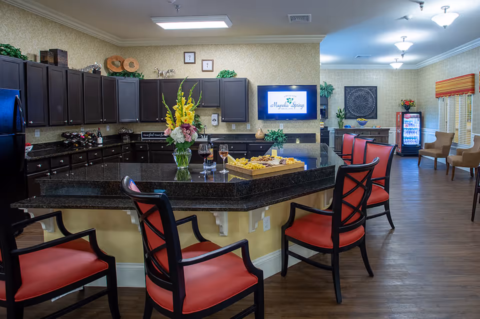 Communal kitchen and lounge with a large dark granite island, red upholstered chairs, cabinetry, and a television displaying the Magnolia Springs logo.