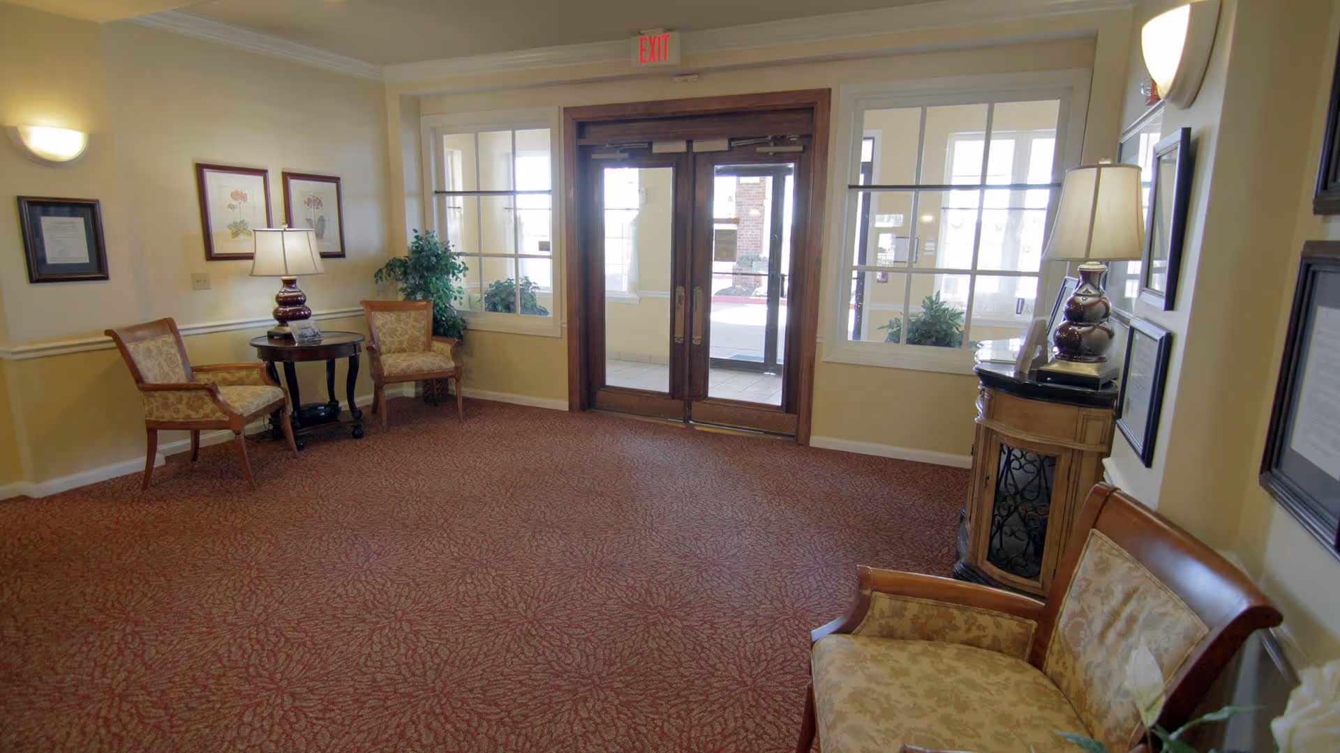 A cozy senior living facility lobby area with patterned carpet, two armchairs and a small round table with a lamp on the left, a wooden double door entrance with glass panels in the center, and a cushioned bench with a lamp on a small cabinet on the right. The walls are decorated with framed pictures and documents, and there are plants near the entrance windows.
