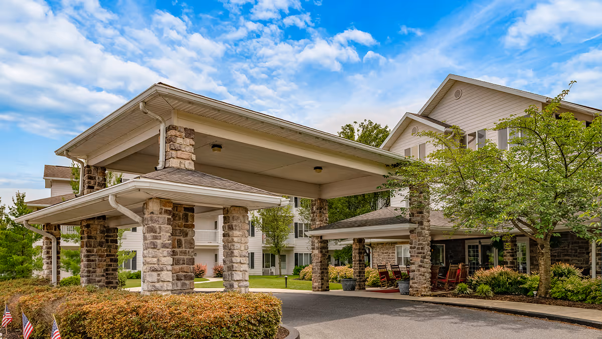 Exterior view of a senior living facility entrance with a covered drop-off area supported by stone pillars, surrounded by greenery and trees under a partly cloudy sky.