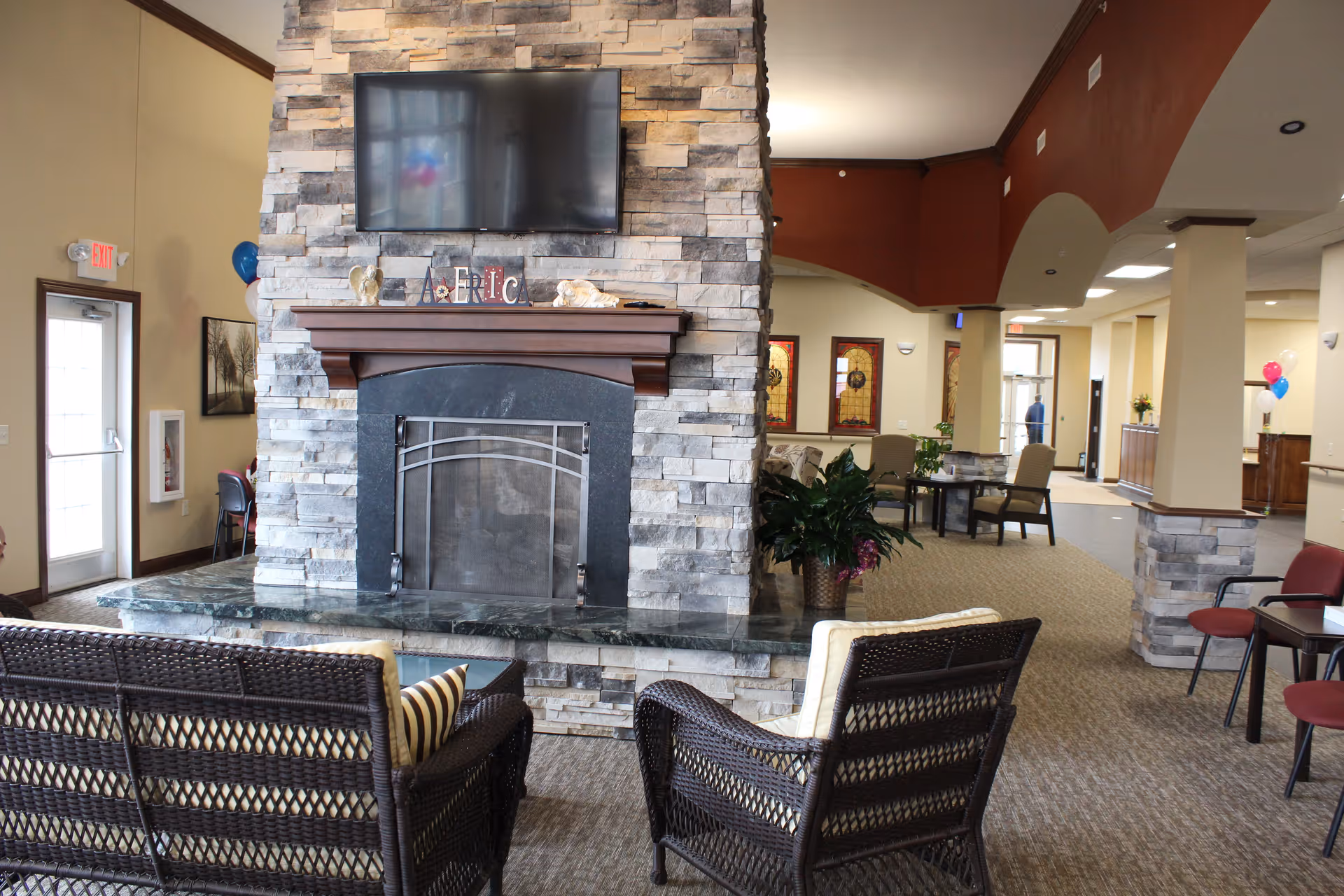 Interior view of a senior living facility lounge area featuring a stone fireplace with a mounted flat-screen TV above it. In front of the fireplace are two wicker chairs with cushions and a small table. The room has carpeted floors, beige walls, and several seating areas with chairs and tables. There are decorative elements like plants, balloons, and framed artwork on the walls. The space is well-lit with natural light coming through glass doors and windows.