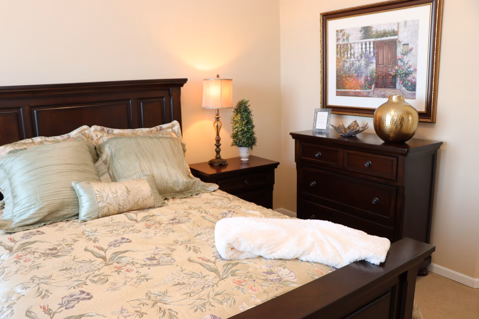 A neatly made bed with floral-patterned bedding and multiple pillows in a bedroom. Next to the bed is a wooden nightstand with a lamp and a small potted plant. Across from the bed is a wooden dresser with a large gold vase, a decorative bowl, and a framed picture on the wall above it.