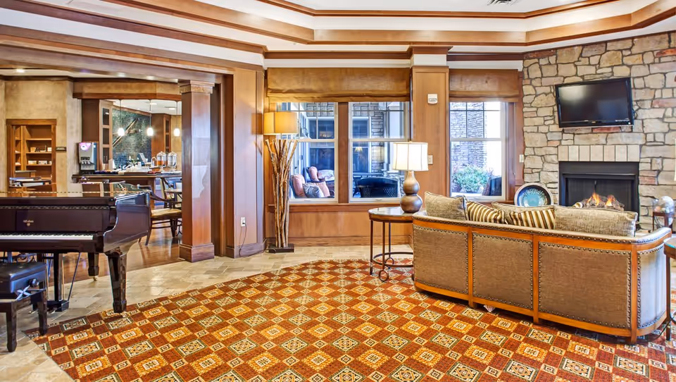 A cozy senior living facility common area with a patterned carpet, a grand piano, a curved sofa with cushions, a stone fireplace with a mounted TV above it, and large windows letting in natural light. There are wooden columns and trim, a floor lamp, and a side table near the sofa. In the background, a dining area with tables and chairs is visible.