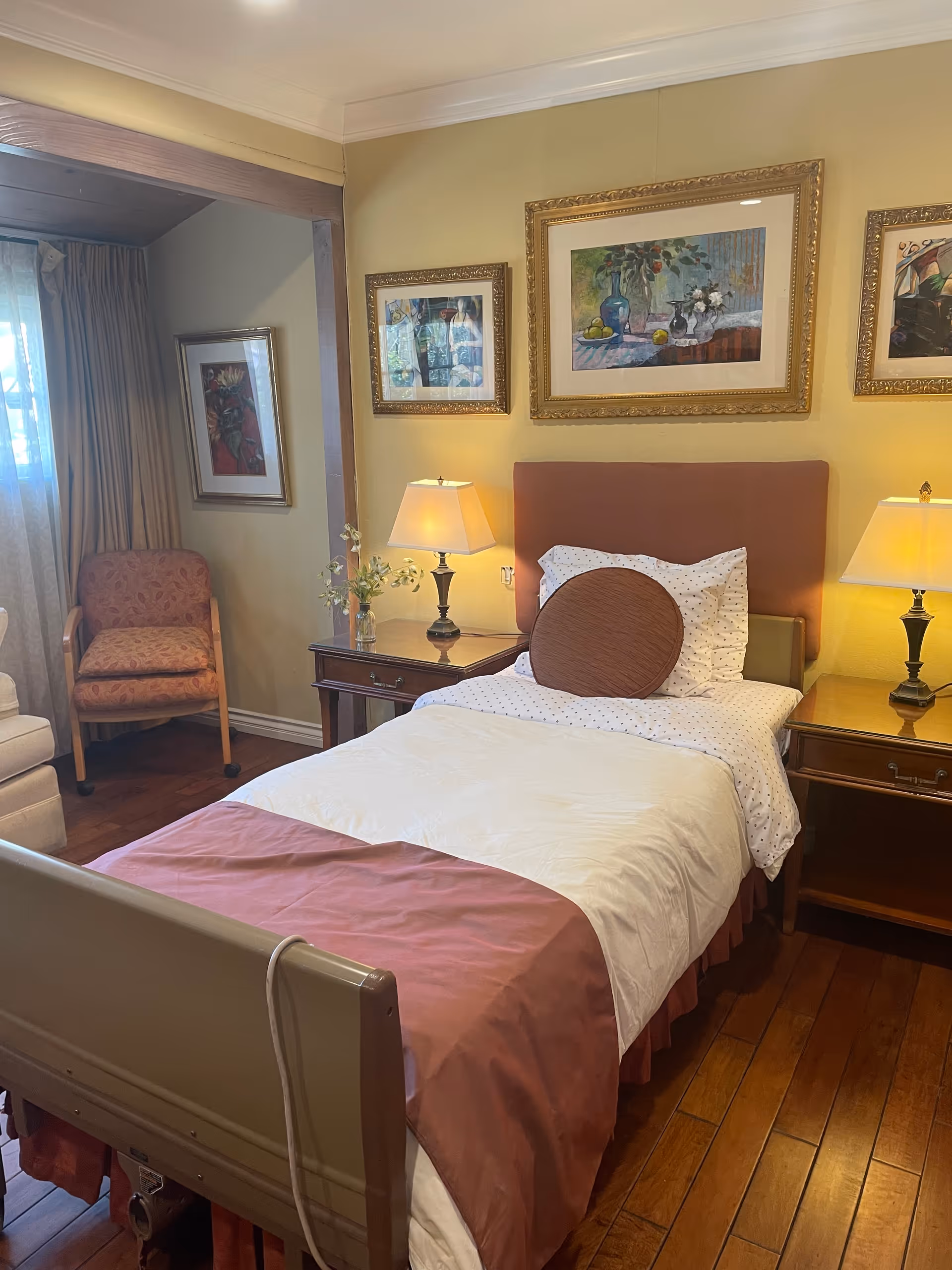 A neatly made single bed with white and pink bedding in a cozy bedroom. The bed has a brown circular pillow and a padded headboard. On each side of the bed are wooden nightstands with table lamps. The walls are decorated with framed paintings. There is a wooden floor and a cushioned armchair near a window with curtains.