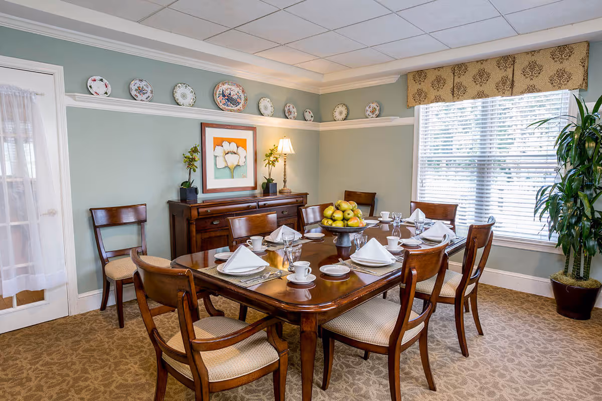 A dining room with a wooden table set for six people, featuring white napkins, cups, saucers, and a centerpiece bowl filled with green apples. The room has light green walls decorated with a row of decorative plates near the ceiling, a framed flower painting, a wooden sideboard with a lamp and plants, a large window with blinds and a patterned valance, and a potted plant in the corner.