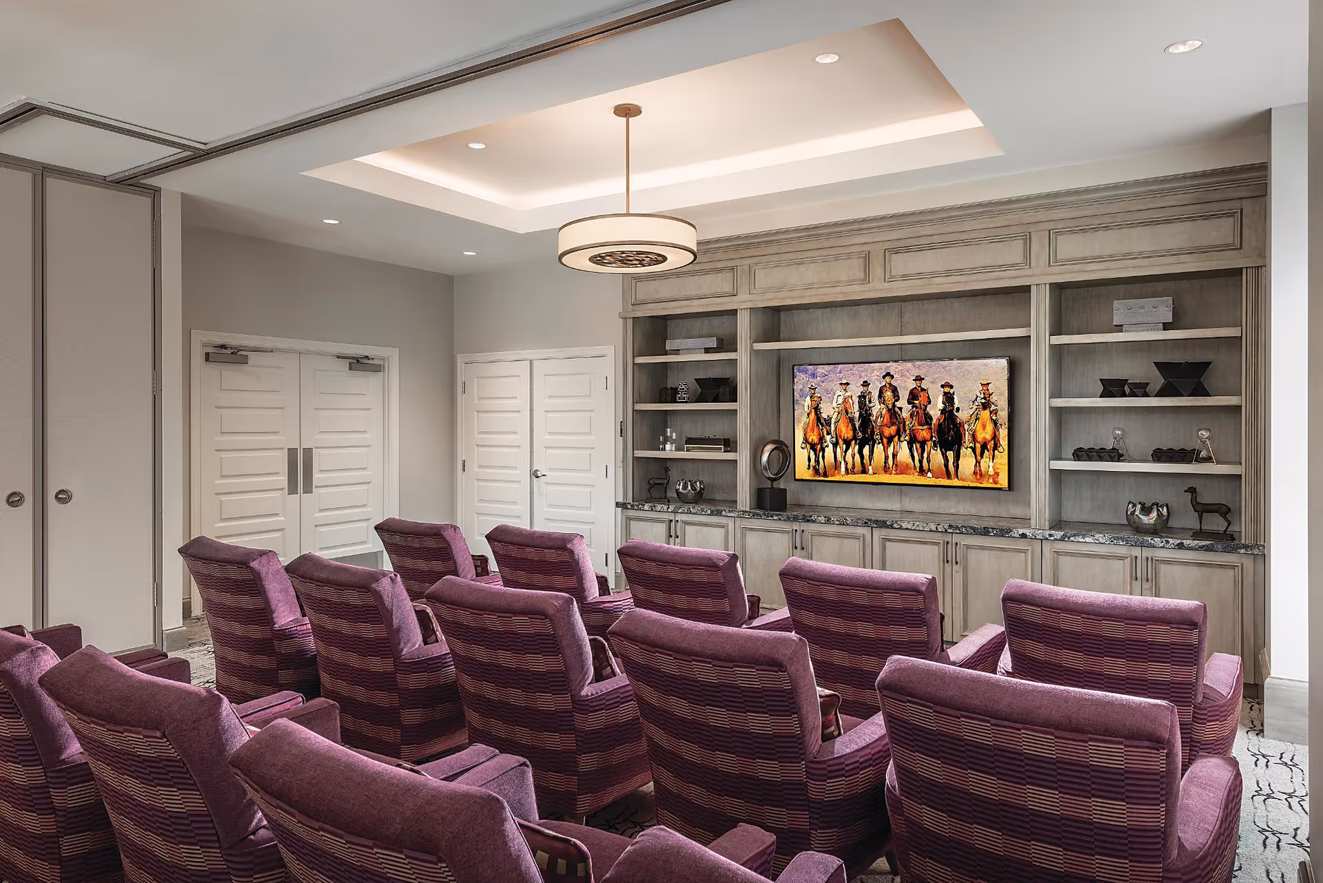 A cozy media room with two rows of purple upholstered chairs facing a large flat-screen TV mounted on a wooden built-in cabinet. The TV displays an image of seven cowboys on horseback. The room has a recessed ceiling light fixture and neutral-colored walls with white double doors on the left side.