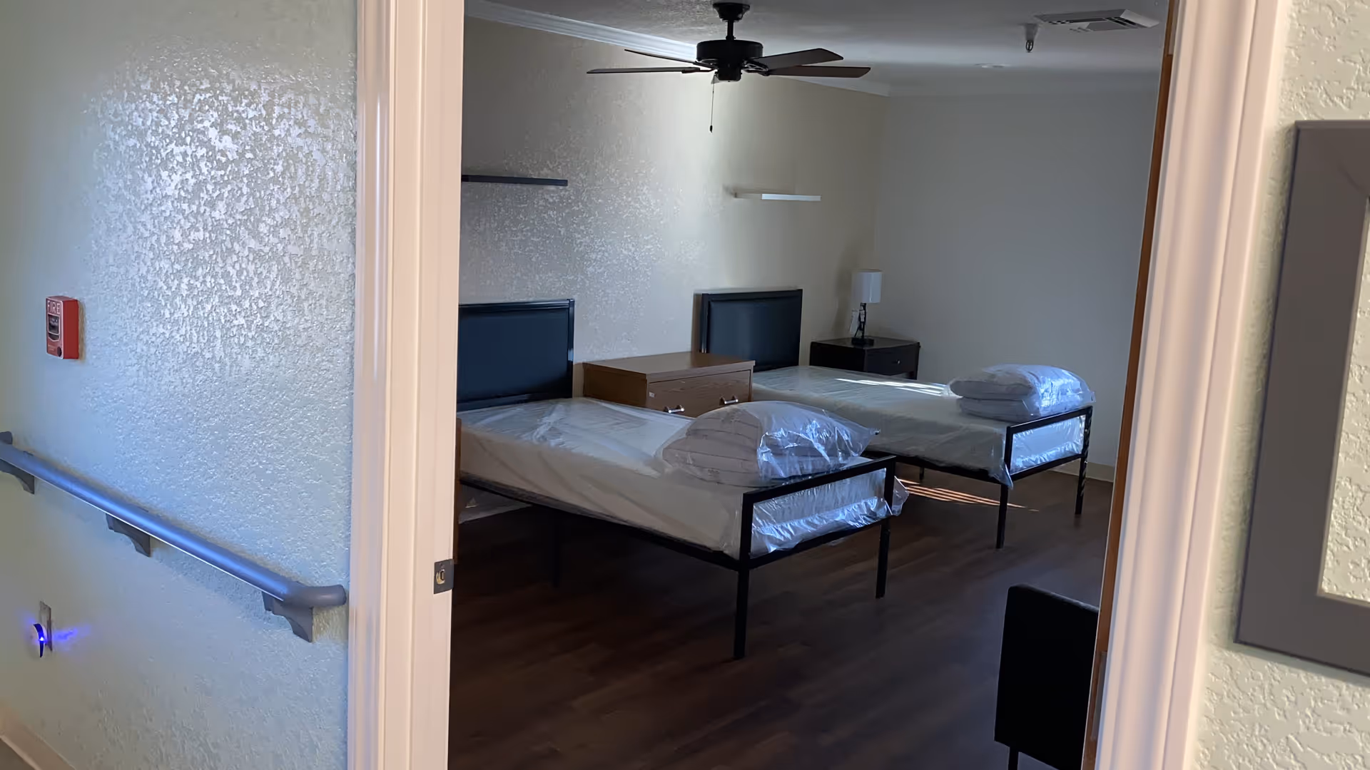 View through an open doorway into a room with two single beds, each with a pillow wrapped in plastic on top. The room has wooden flooring, a ceiling fan, a nightstand with a lamp, and a small dresser between the beds. The hallway outside the room has a handrail and a fire alarm on the wall.