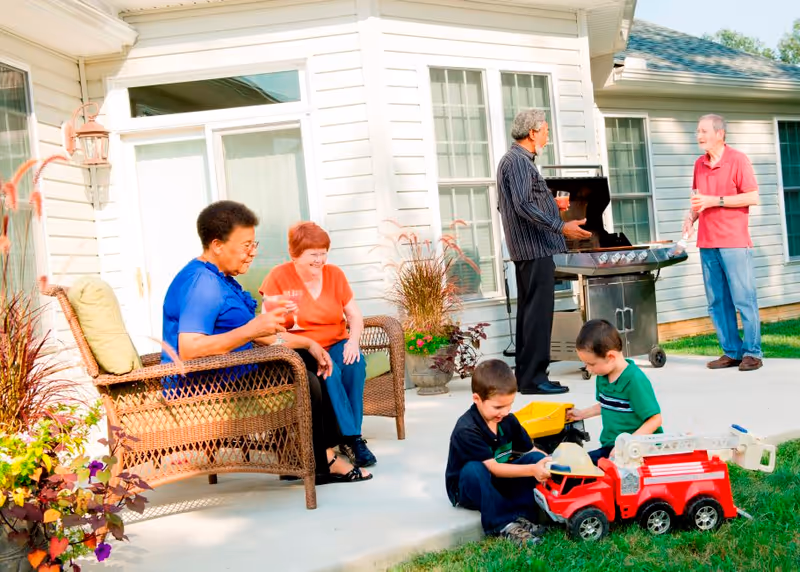 Two elderly women sitting on wicker chairs on a patio outside a house, holding drinks and smiling. Two elderly men stand near a barbecue grill, also holding drinks and conversing. Two young boys are playing with toy trucks on the grass near the patio.