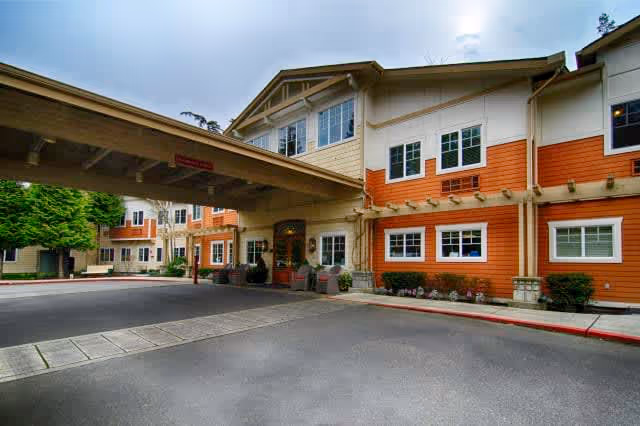 Exterior view of a senior living facility named Aegis Lodge, showing a covered entrance with a driveway, multiple windows, and a combination of beige and orange siding on the building.