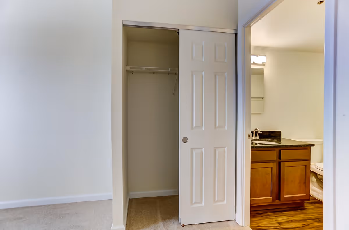 Interior view showing a partially open closet with a white sliding door and an adjacent bathroom with a wooden vanity, black countertop, sink, mirror, and toilet. The floor in the closet area is carpeted, while the bathroom has wood-style flooring.